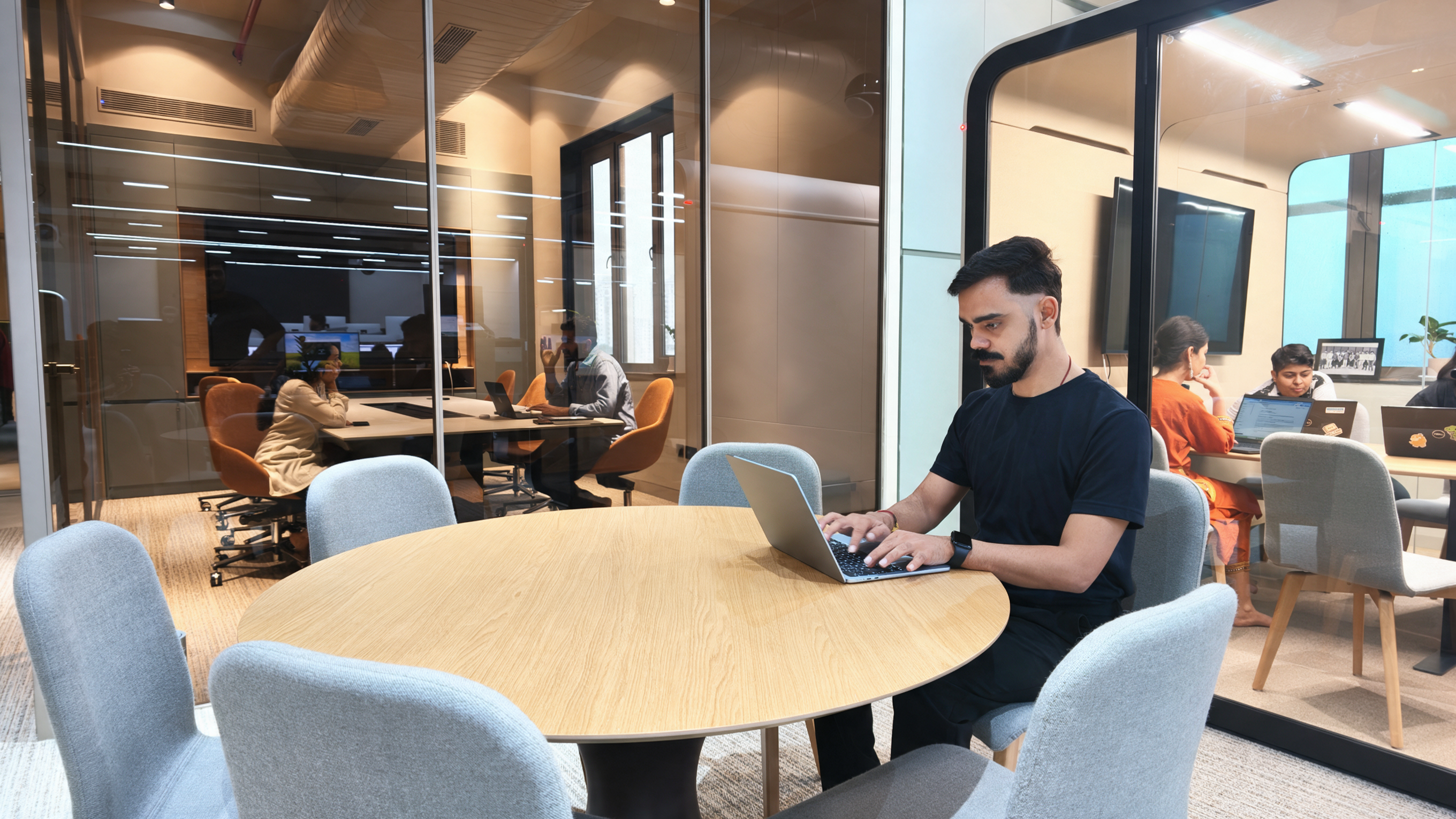 A MoreTasks colleague working on a laptop in the Noida office, with the wider team at desks behind the glass